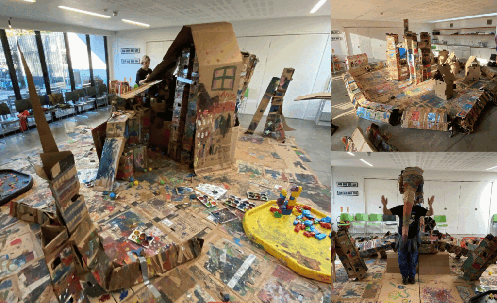 A large indoor play space filled with colourful cardboard structures, painted tunnels, and art materials, created collaboratively by children.